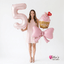 Woman in a pink dress holding a large pink number 5 balloon and a cupcake with a bow, against a white background.