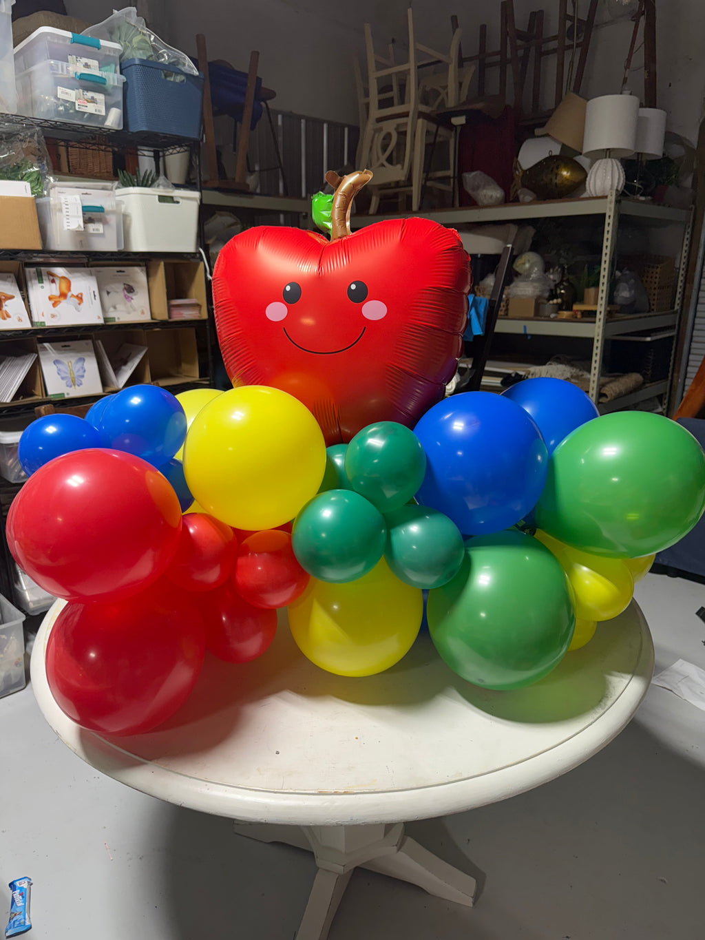 Colorful balloon sculpture shaped like a caterpillar with a large red apple balloon on top, in a cluttered room.