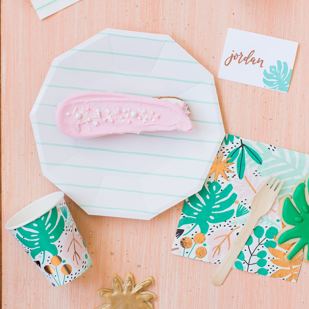 Party supplies including a pink cookie, striped paper plate, and tropical-themed cups and napkins on a wooden surface.