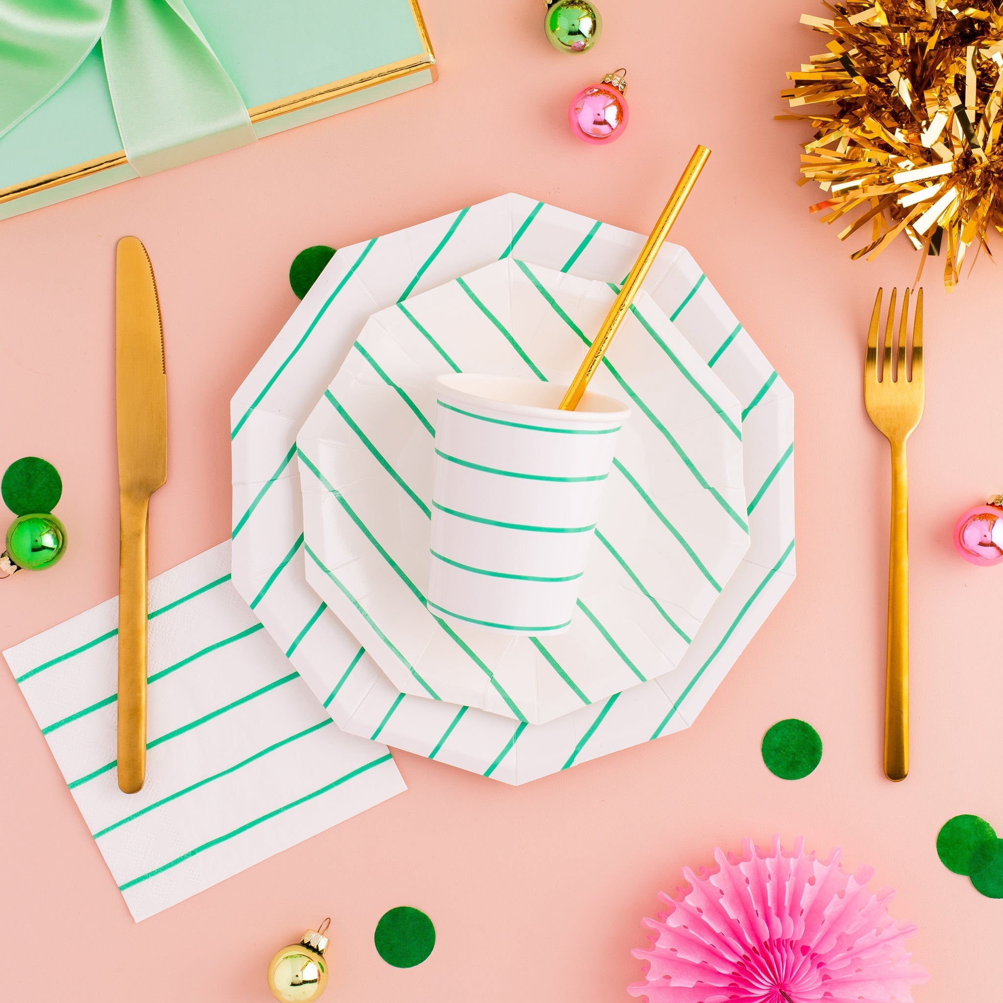 Hexagonal paper plates with green stripes on a pink surface with gold cutlery and festive decorations.