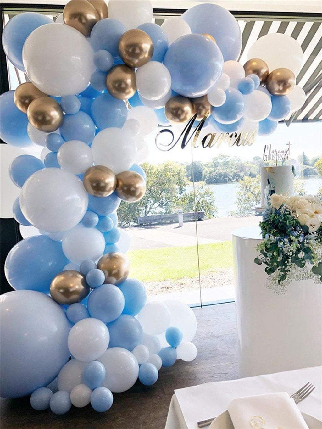 Balloon arch with blue, white, and gold balloons in a room with a table and flowers.