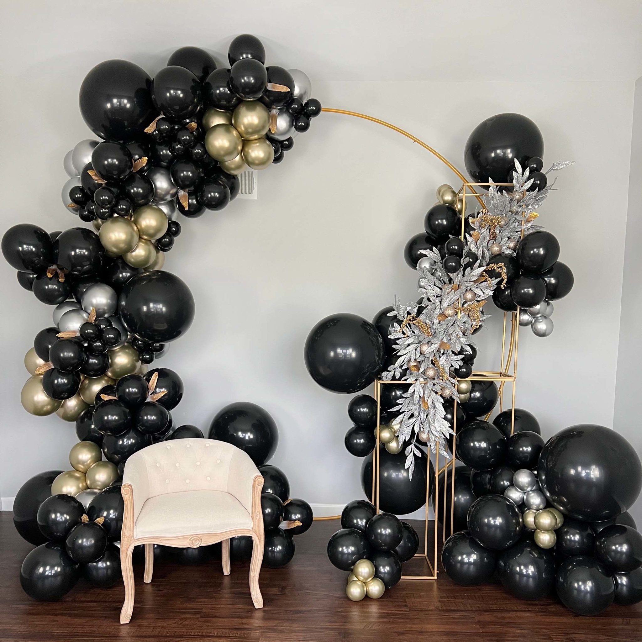 Balloon arch with black, gold, and silver balloons in a room with a chair. Main image