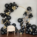 Balloon arch with black, gold, and silver balloons in a room with a chair.