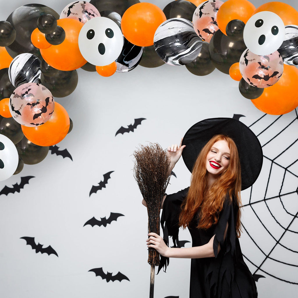 Woman in a Halloween costume with a broom, surrounded by balloons and decorations on a white wall.