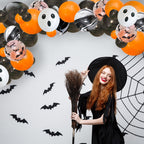 Woman in a Halloween costume with a broom, surrounded by balloons and decorations on a white wall.