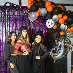 Three women posing in front of a Halloween-themed balloon arch with purple tinsel curtain.