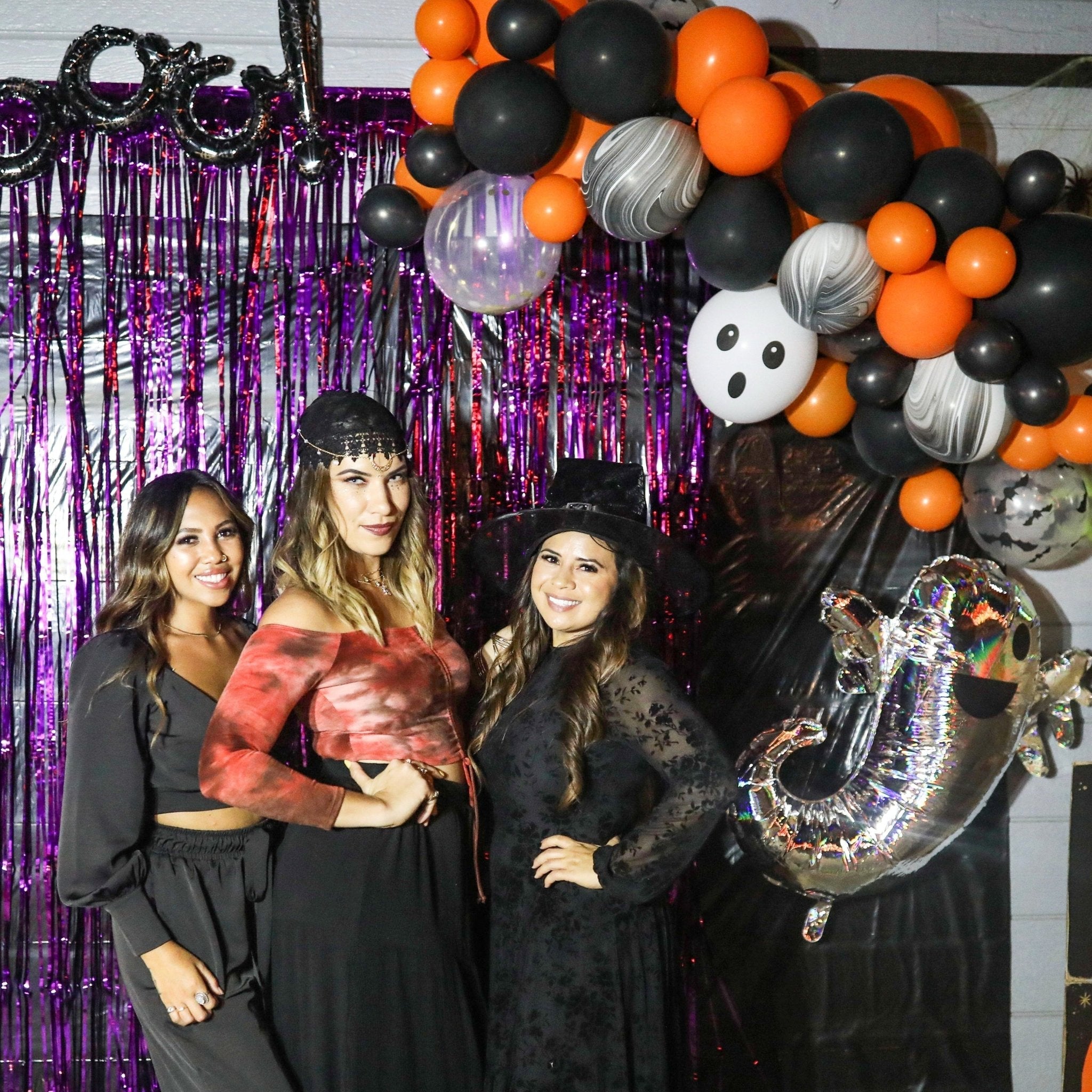 Three women posing in front of a Halloween-themed balloon arch with purple tinsel curtain.