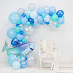 Balloon arch with blue, green, and white balloons and a shark balloon in front of a white chair.