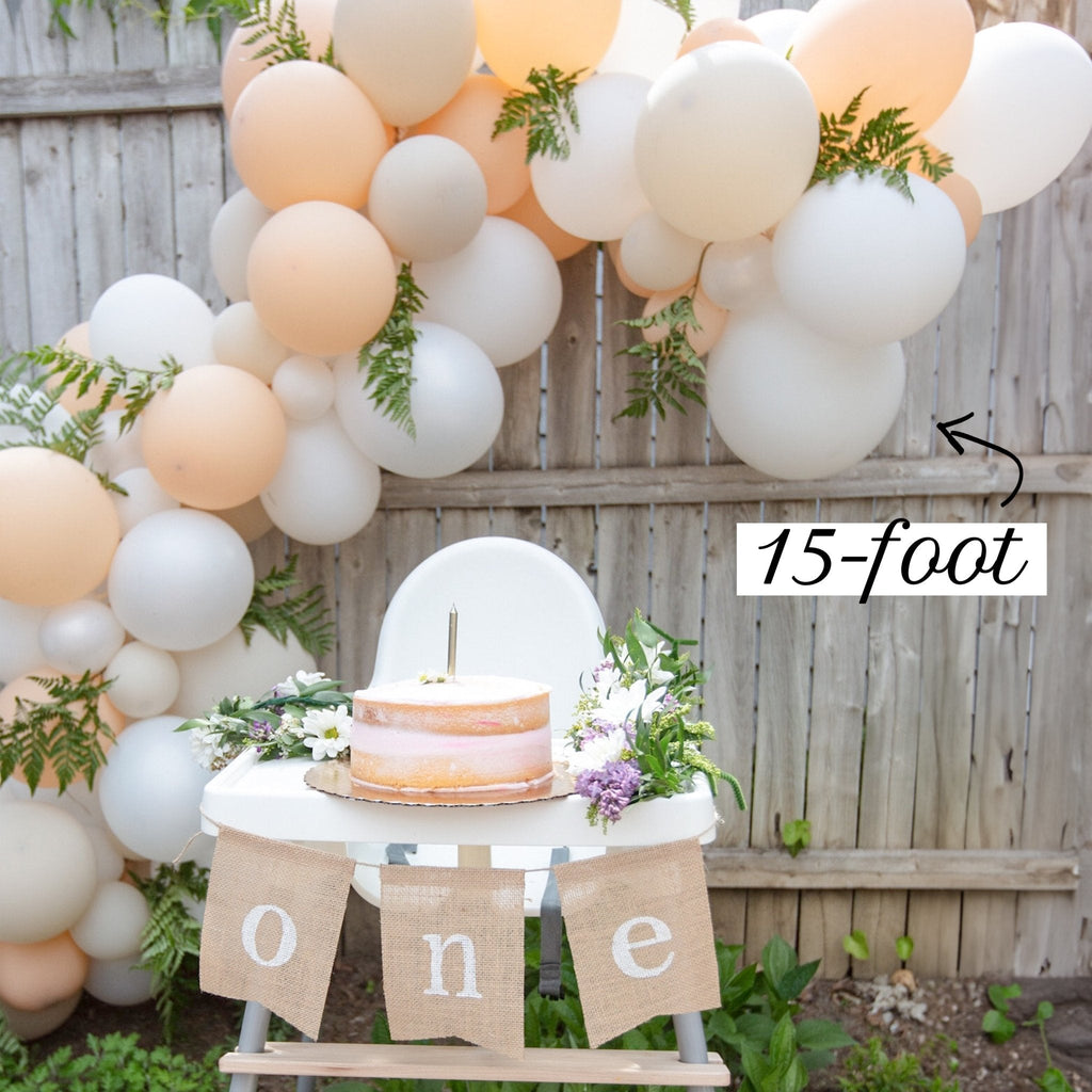 Balloon Garland with peach, white, and green balloons, a cake on a table, and 'one' banner in front of a wooden fence.