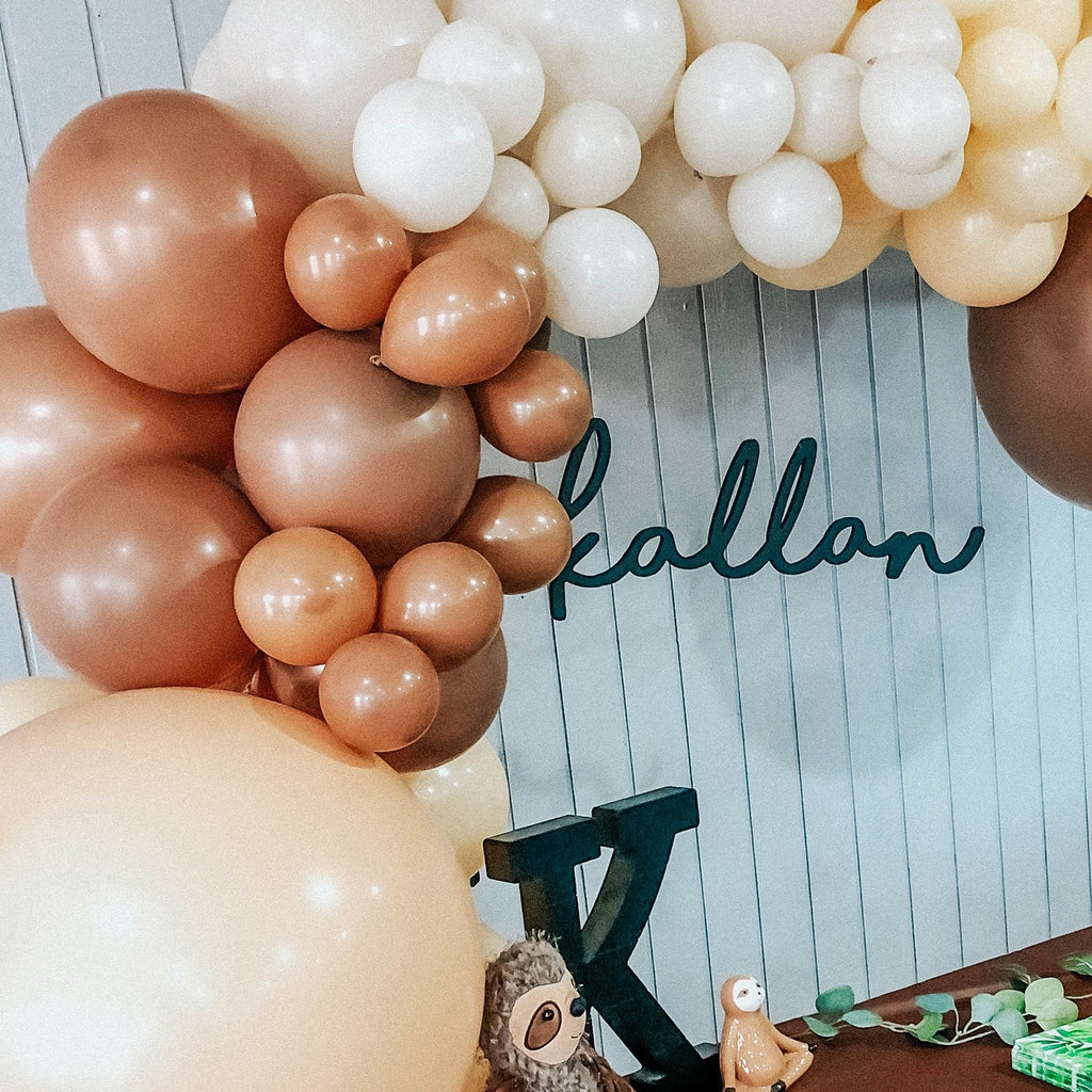 Balloon arch with brown, white, and gold balloons against a wooden background with 'kallan' written on it.