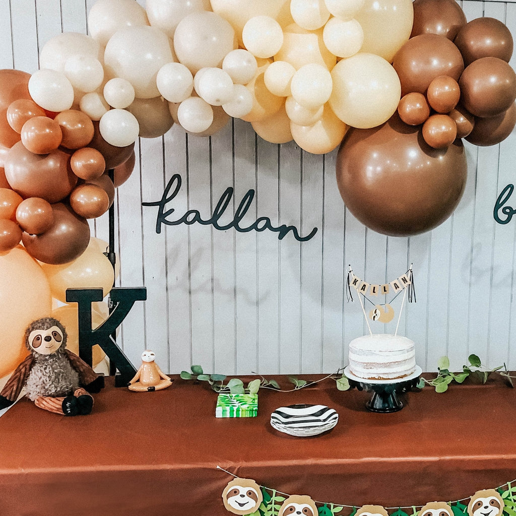 Decorative setup with balloons, sloth toys, and a cake on a table against a wooden wall.