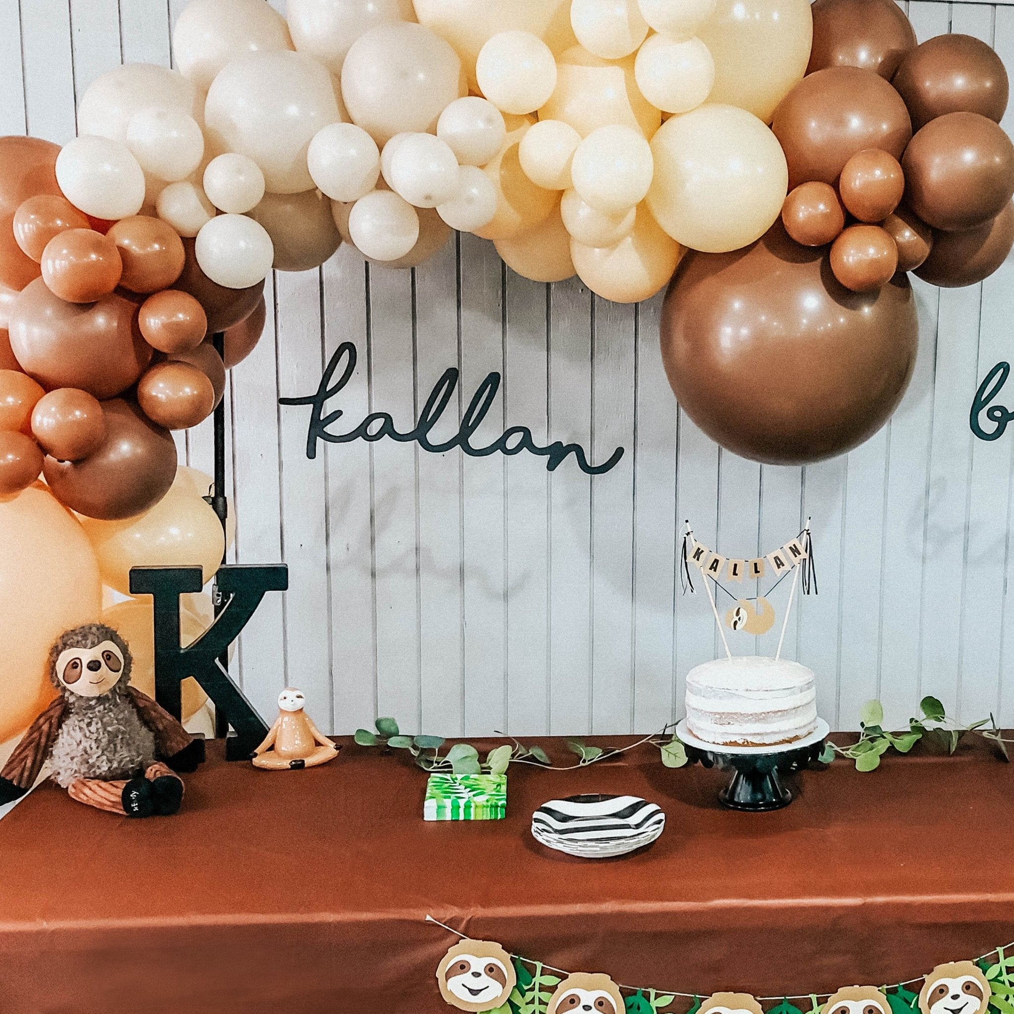Decorative setup with balloons, sloth toys, and a cake on a table against a wooden wall.