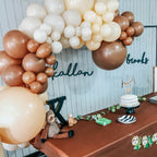 Balloon arch with brown, beige, and white balloons above a table with decorations and a name sign.