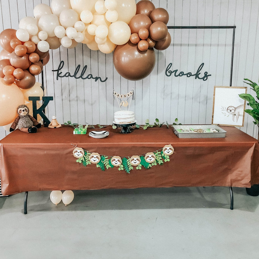 Decorated table with balloons, cake, and name signs against a wooden wall.