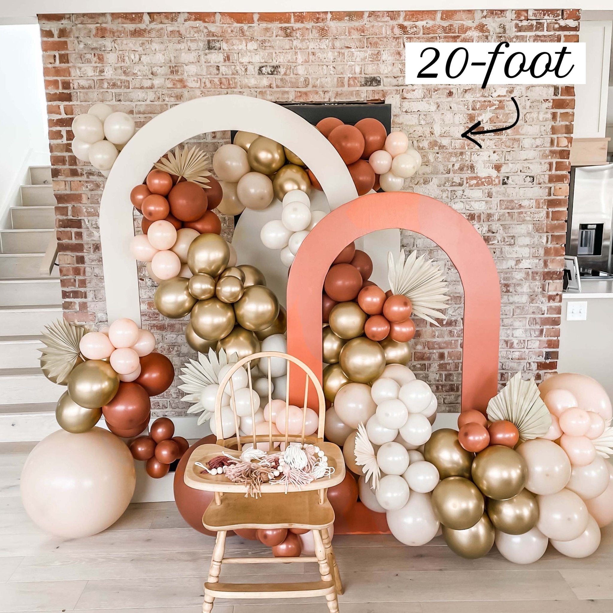 Balloon arches in pink, gold, and white with a chair underneath against a brick wall.