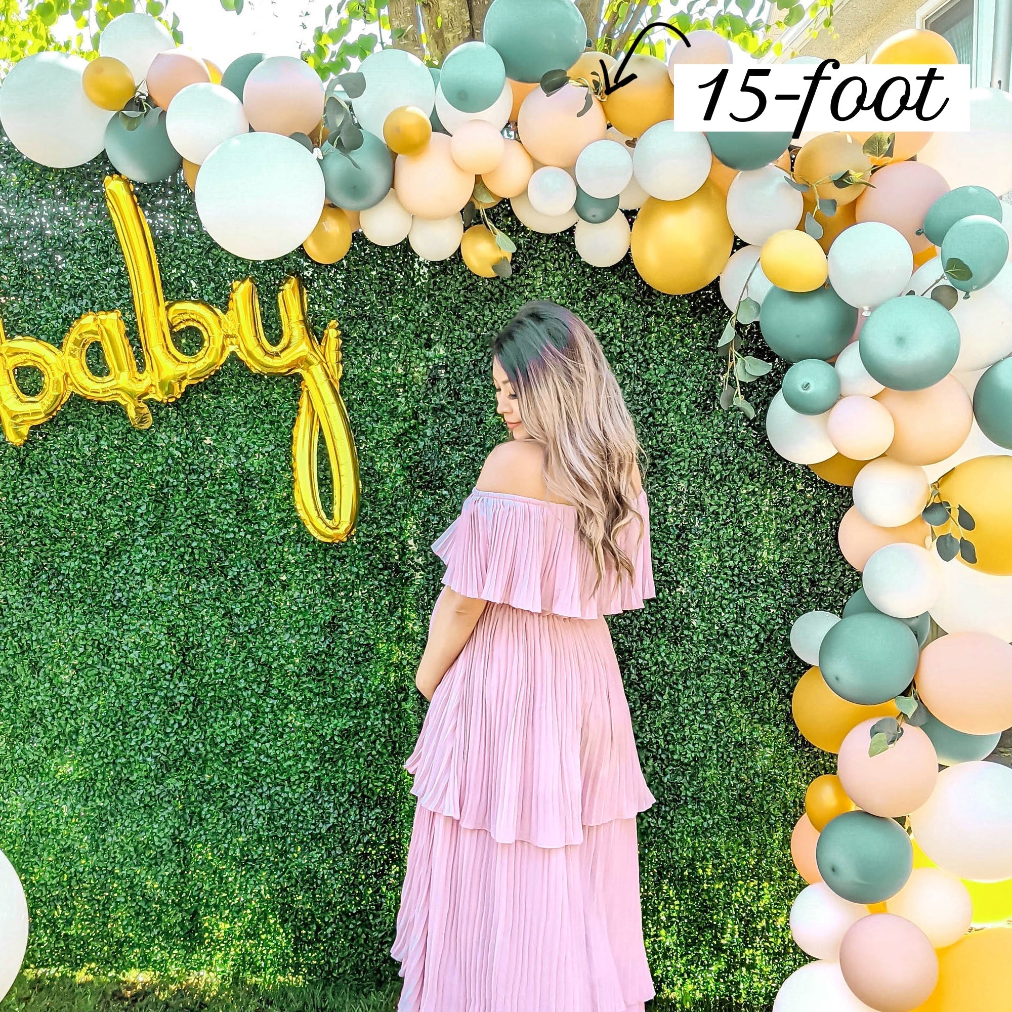Woman in a pink dress standing under a large balloon arch with 'baby' balloons and a 15-foot label.