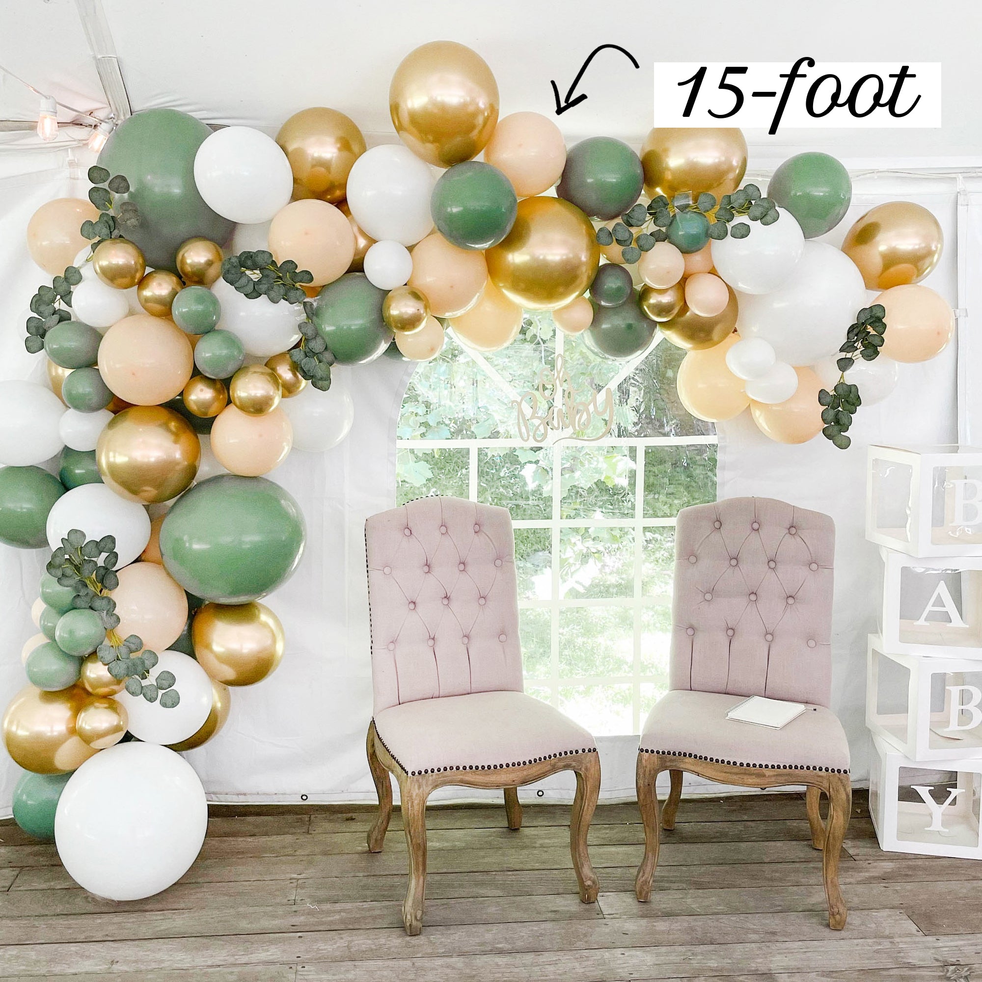 Balloon arch with green, gold, and white balloons in a room with two chairs.