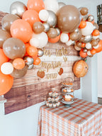 Decorative balloon arch with orange, white, and brown balloons above a dessert table with a plaid tablecloth.