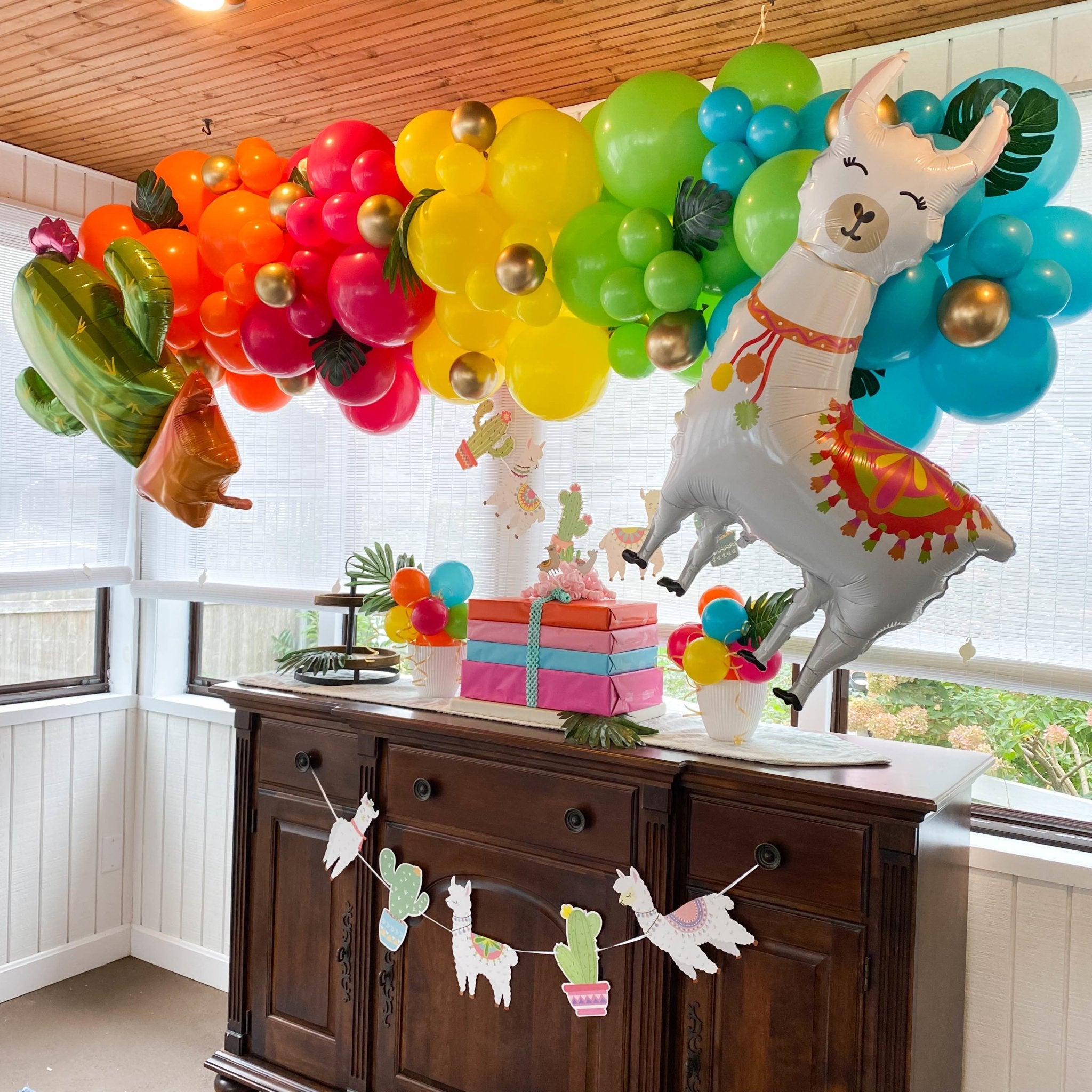 Decorative setup with colorful balloons, a llama balloon, and a wooden cabinet with party decorations. Main image