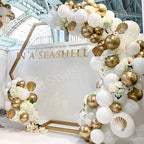 Balloon arch with gold and white balloons and shell decorations in front of a branded wall.