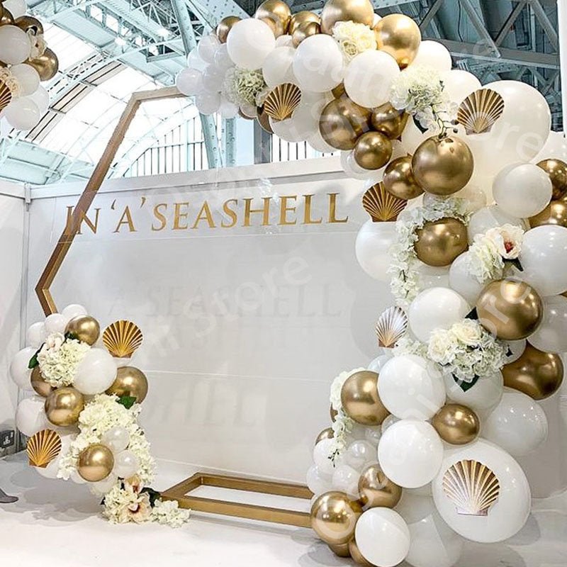 Balloon arch with gold and white balloons and shell decorations in front of a branded wall.