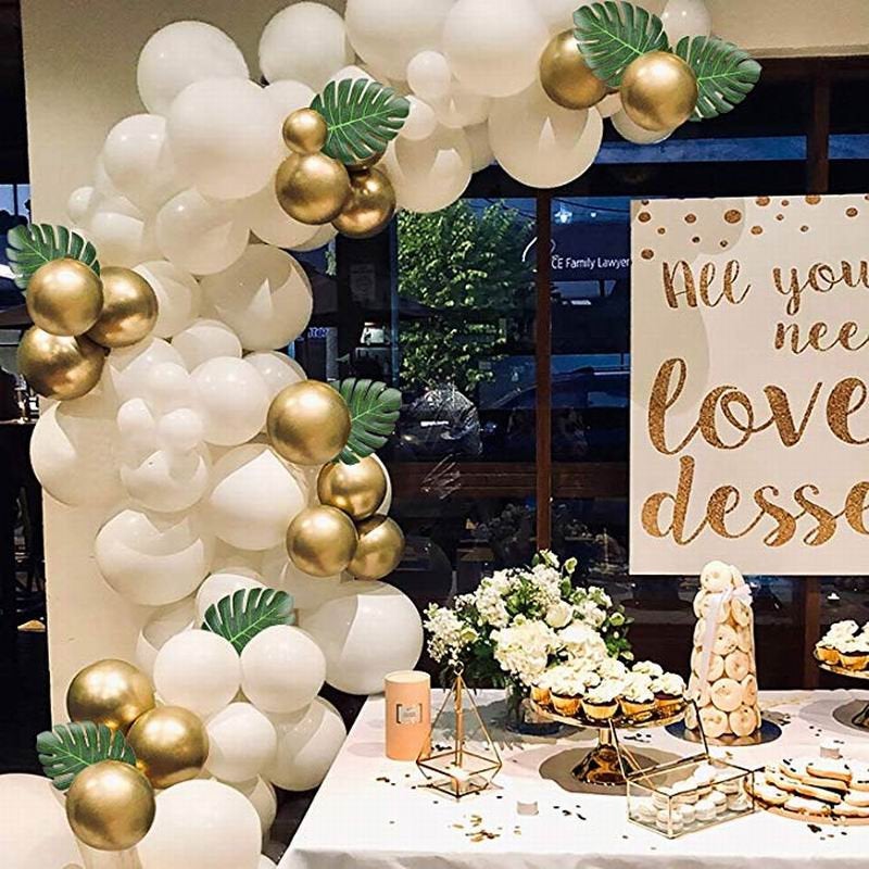 Decorative balloon arch with gold and white balloons, green leaves, and a dessert table in the background.