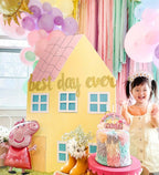 Child in a party setting with a 'best day ever' house, balloons, and a cake.