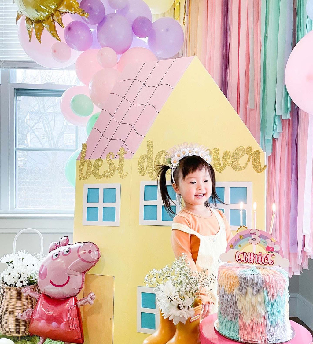 Child in a decorated room with balloons, a house-shaped backdrop, and a birthday cake.