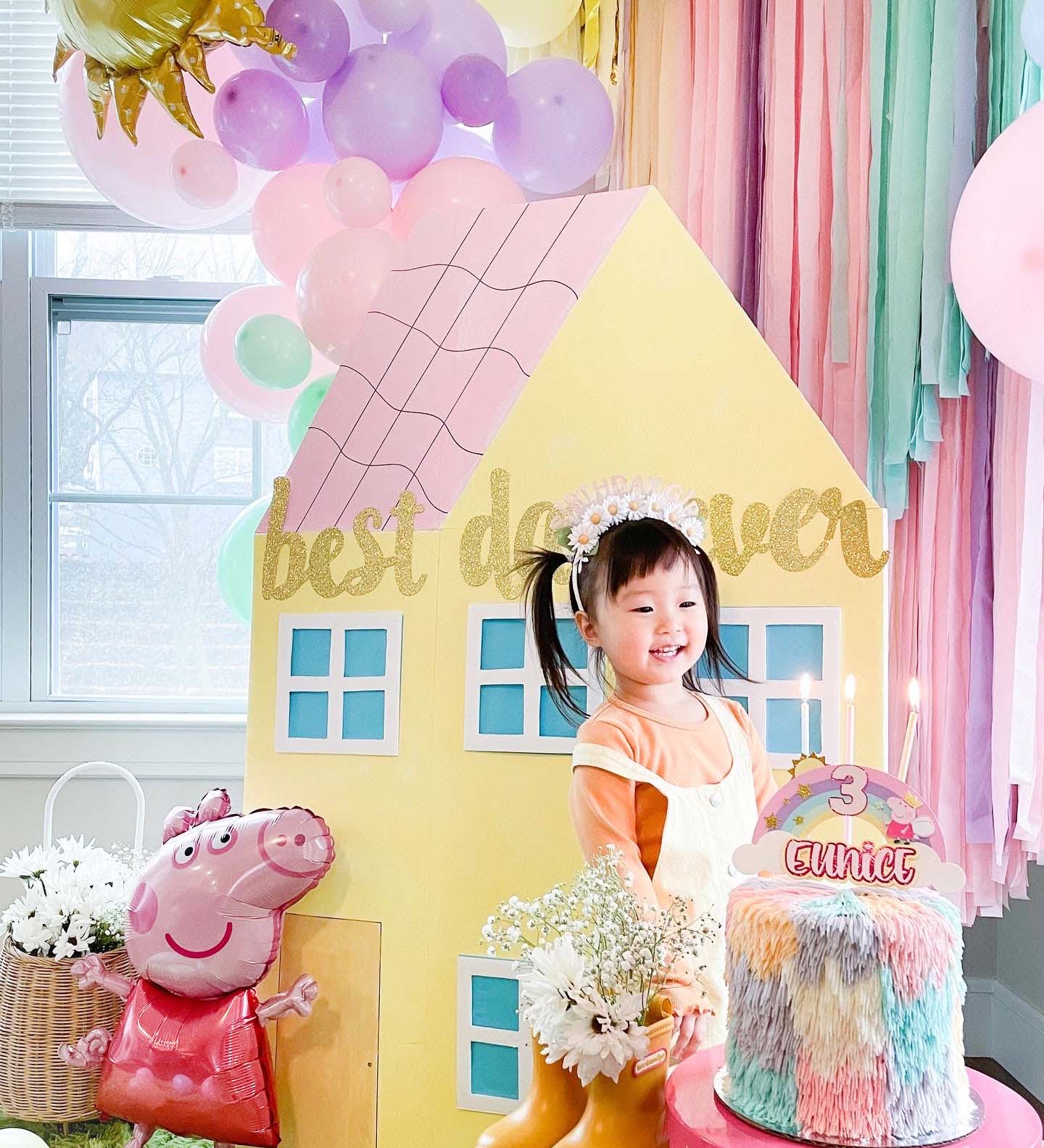 Child in a decorated room with balloons, a house-shaped backdrop, and a birthday cake.