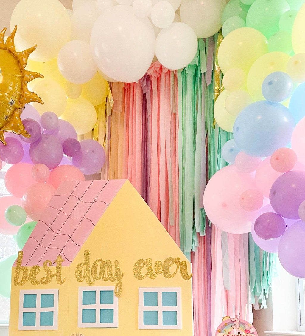 Decorative setup with colorful balloons, pastel curtains, and a house-shaped sign reading 'best day ever'.