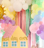 Decorative setup with colorful balloons, pastel curtains, and a house-shaped sign reading 'best day ever'.