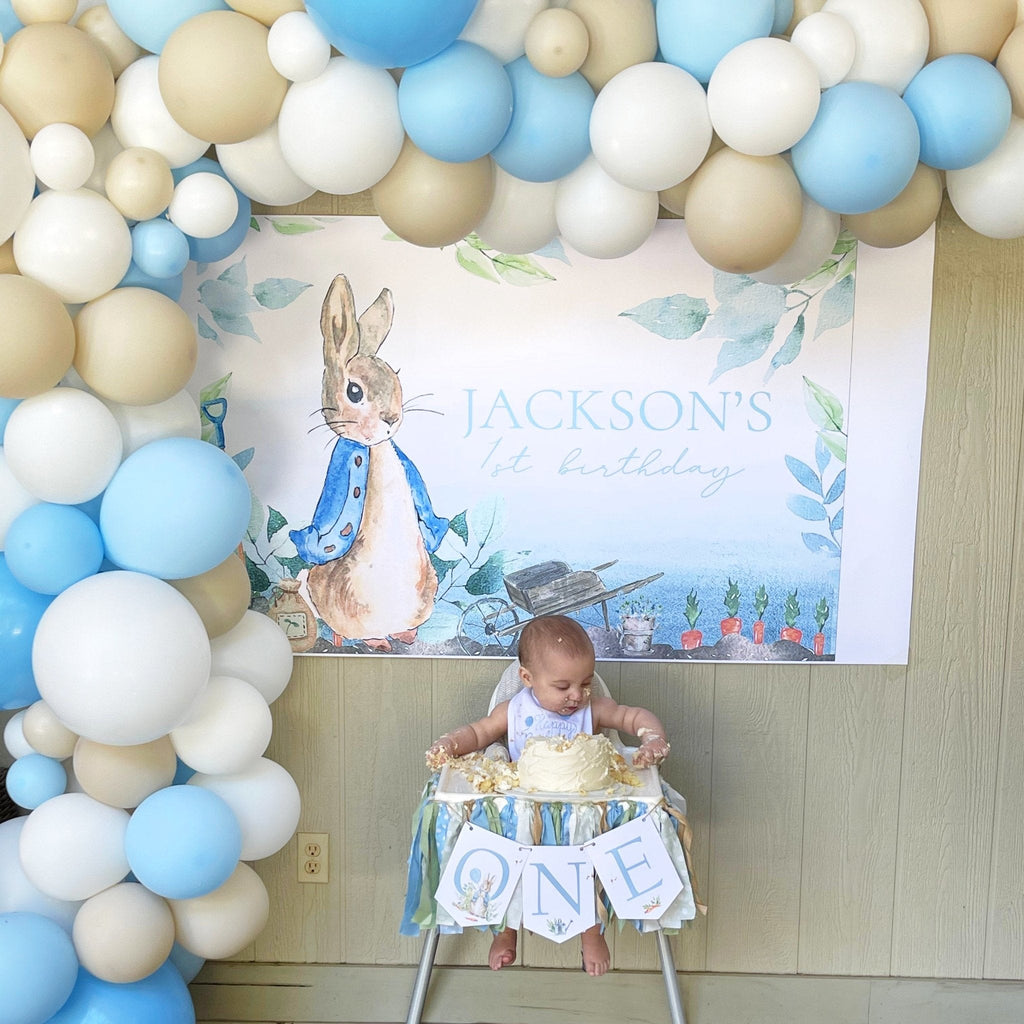 Baby celebrating a first birthday with a balloon arch and 'Peter Rabbit' themed backdrop.