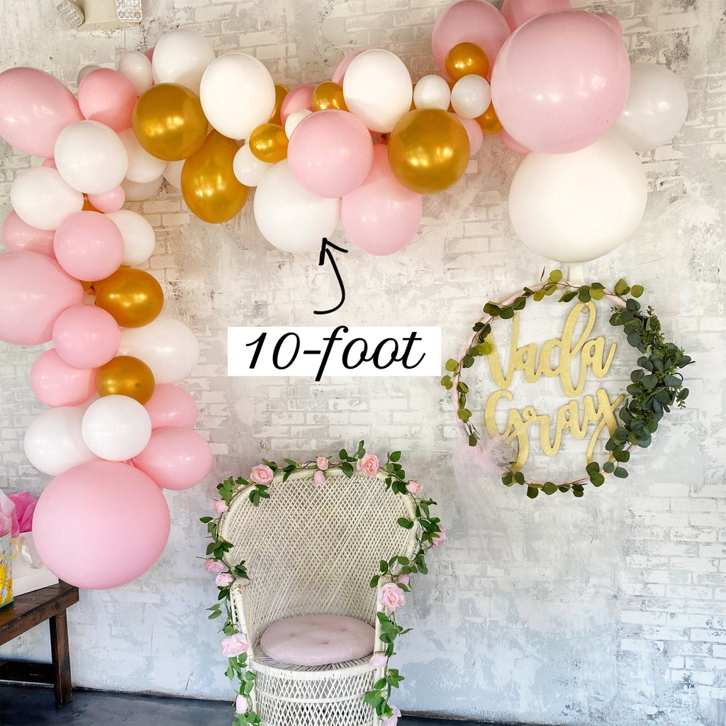 Balloon arch with pink, gold, and white balloons against a textured wall with a chair and decorative wreath.
