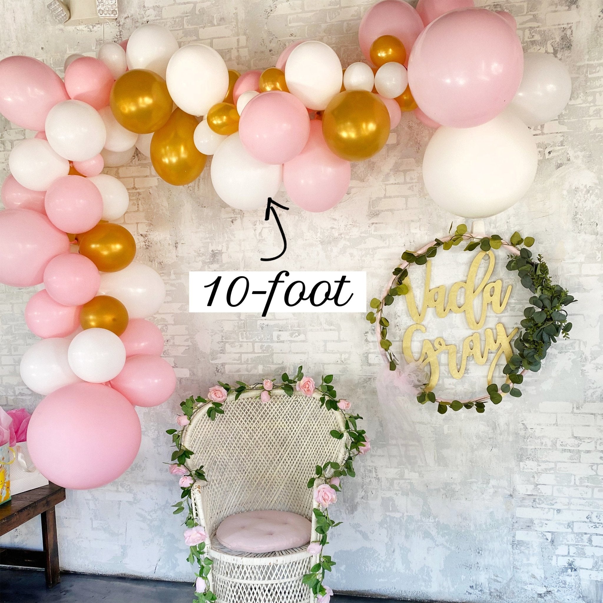 Balloon arch with pink, gold, and white balloons against a textured wall with a chair and decorative wreath.