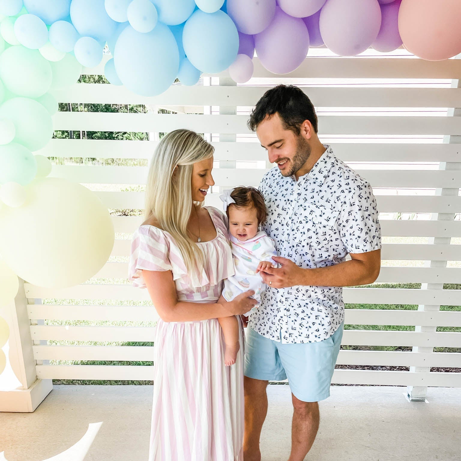 Family of three with a baby standing under colorful balloons in a bright room.