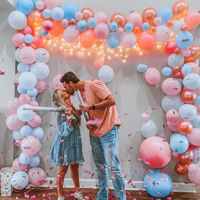 Couple standing under a colorful balloon arch with confetti on a light-colored wall.