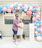 Couple standing in front of a balloon arch with pink, blue, and white balloons, celebrating a baby shower.