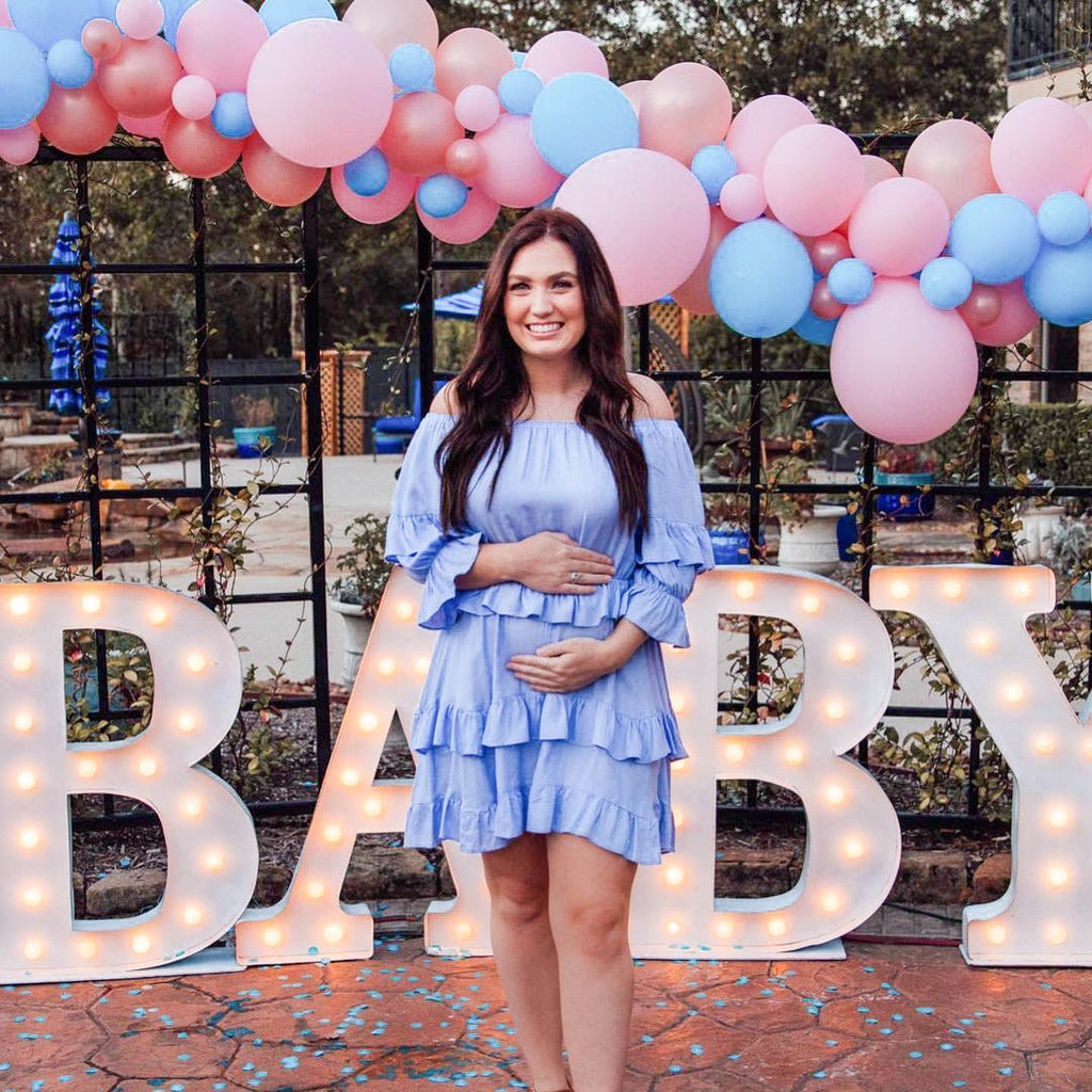 Pregnant woman in a light blue dress standing in front of a 'BABY' sign with balloons at a baby shower.