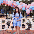 Pregnant woman in a light blue dress standing in front of a 'BABY' sign with balloons at a baby shower.