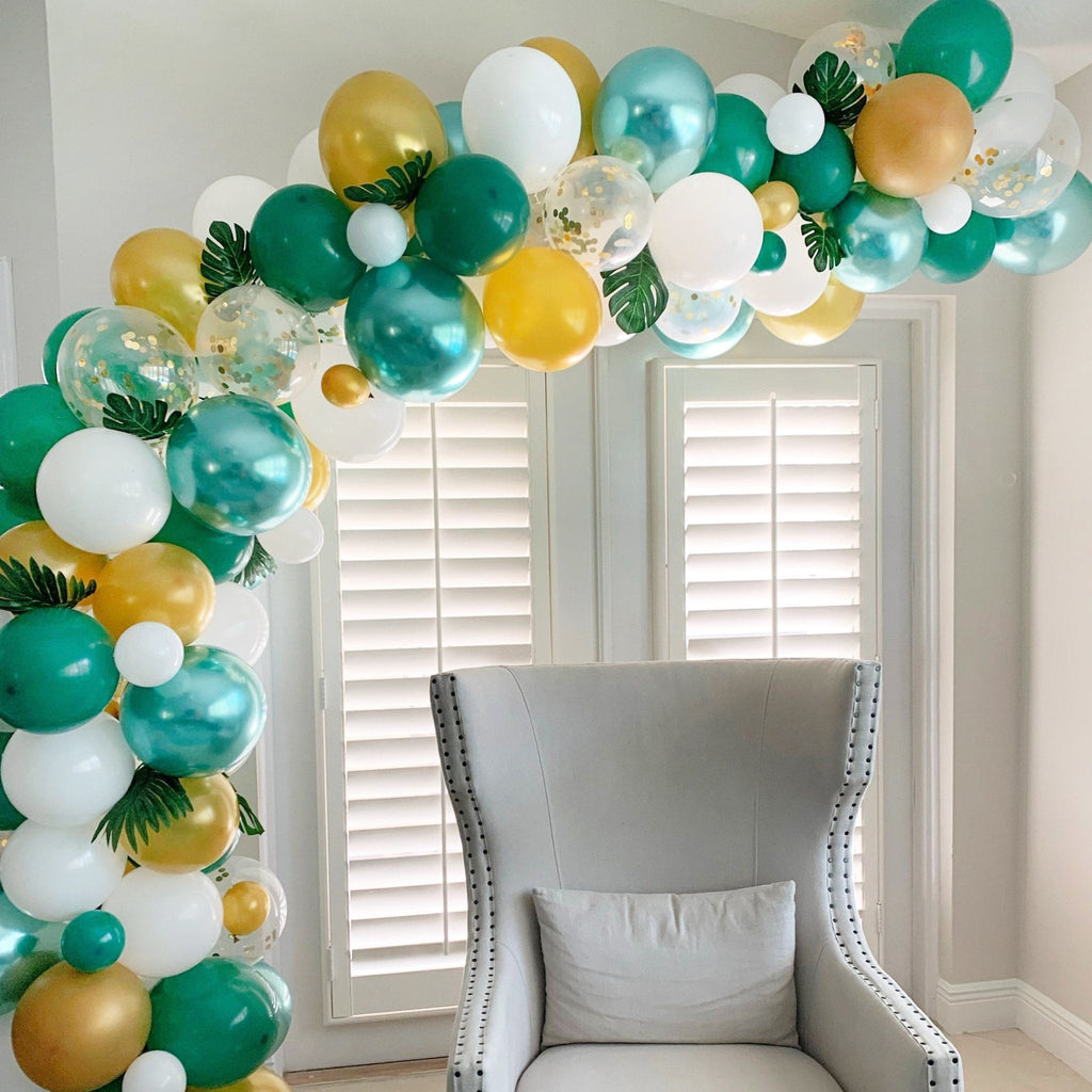 Balloon arch with green, gold, and white balloons in a room with a chair and window blinds.