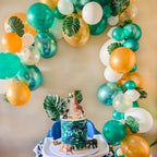 Decorative balloon arch with green, gold, and white balloons, featuring a cake with animal figurines and leaf patterns.