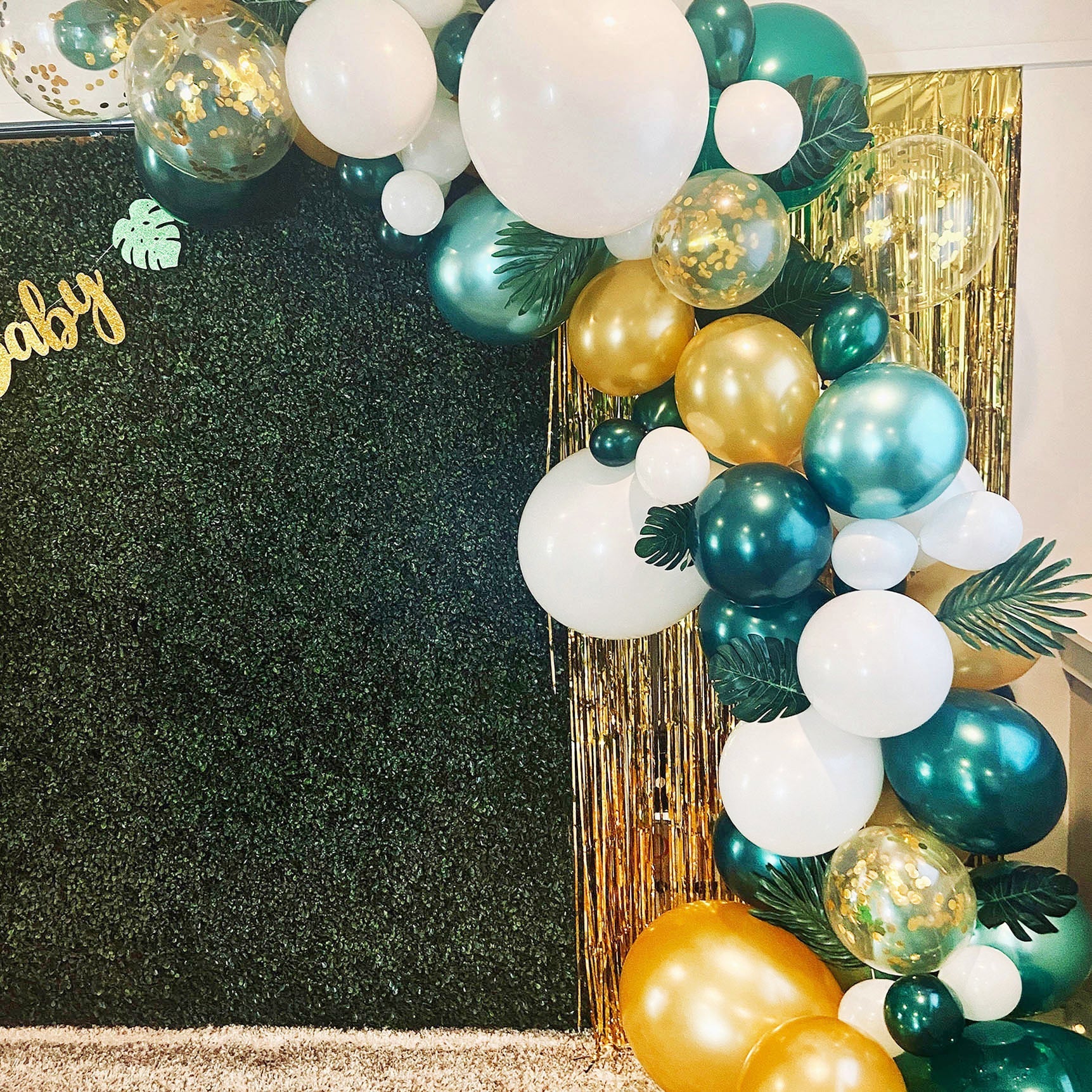 Decorative balloon arch with green, gold, and white balloons against a textured black wall.