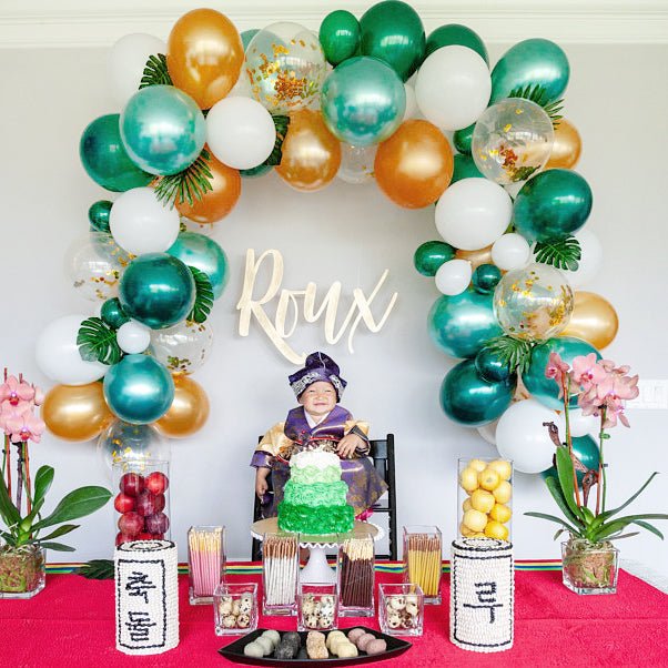Decorative balloon arch with 'Roux' sign above a table with food and plants.