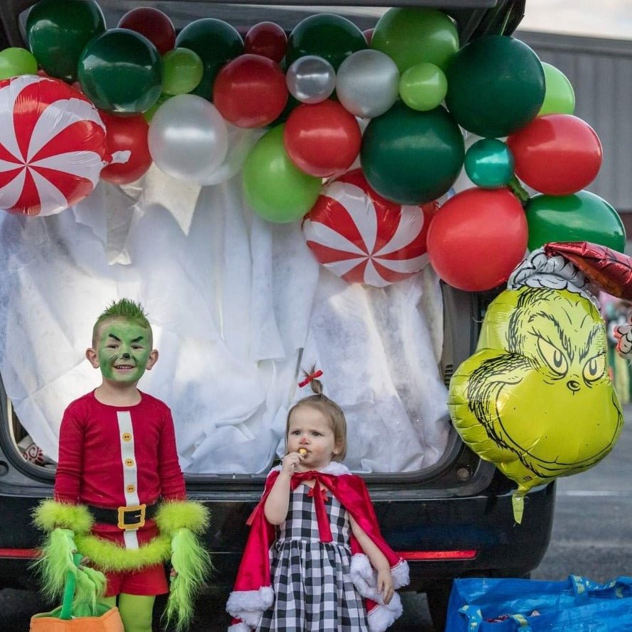 Two children in festive costumes sitting in a decorated car trunk with balloons and decorations.