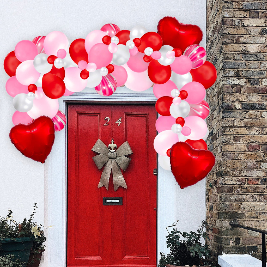 Decorative balloon arch with red, pink, and white balloons above a red door with a gold bow.