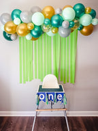 High chair with 'one' banner under a balloon arch with green, gold, and white balloons against a plain wall.