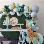 Birthday party setup with balloon arch, cake, and decorations against a green wall.