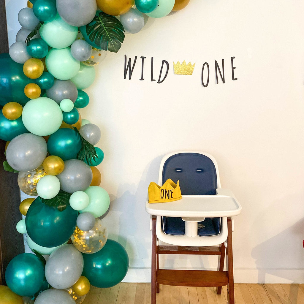 High chair with 'Wild One' crown decoration in front of a balloon arch with 'Wild One' text on a wall.